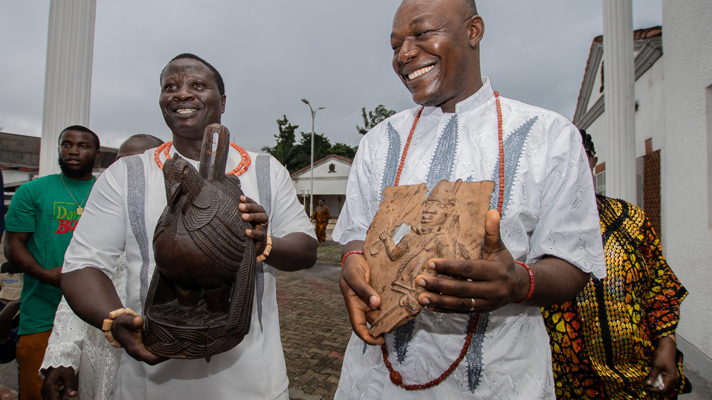 Two men wearing white, holding art objects and smiling