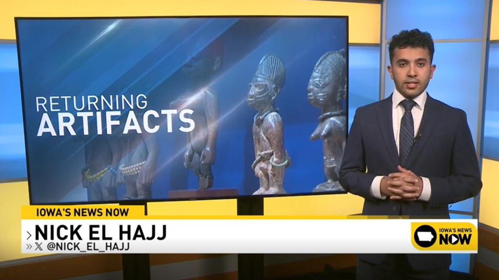 A person in a suit presenting the news standing next to a tv screen that reads "Returning Artifacts" 