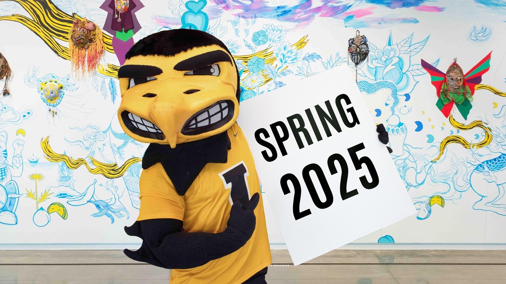 A black and yellow hawk mascot, Herky, holding up a sign that reads "Spring 2025"