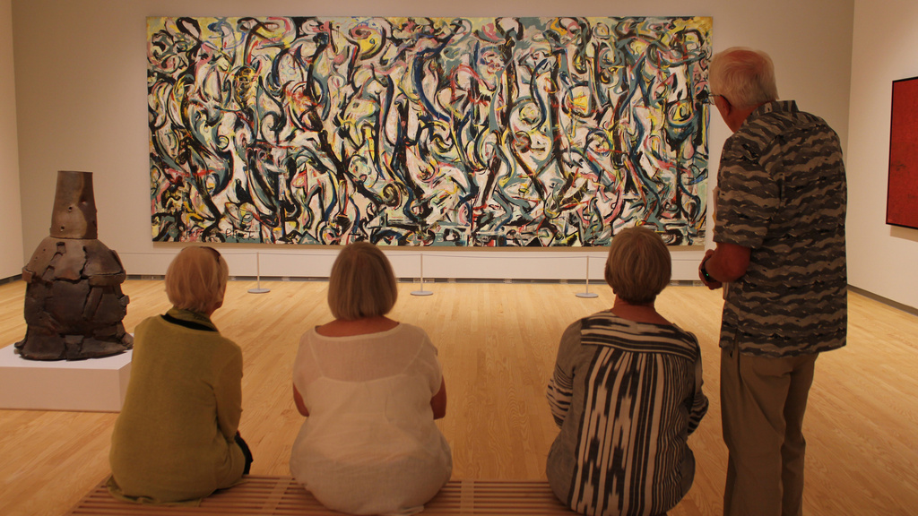 Four people observe an abstract painting in a gallery. One person stands while three sit on a bench. Nearby is a metal sculpture on display.
