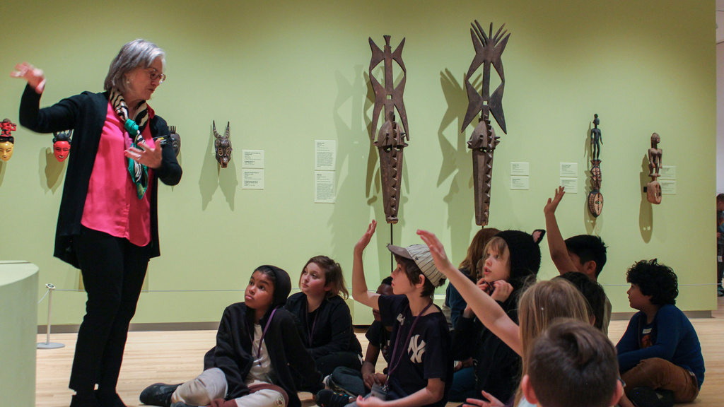A woman speaks to a group of children sitting on the floor in a museum. Some children have their hands raised. Masks and sculptures are displayed on the green wall behind them.