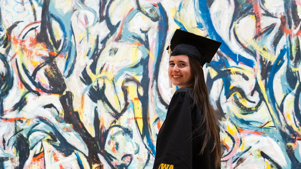 Gallery Host and Campus Engagement Coordinator Annelies Knight poses in her graduation gown in front of Jackson Pollock's Mural.