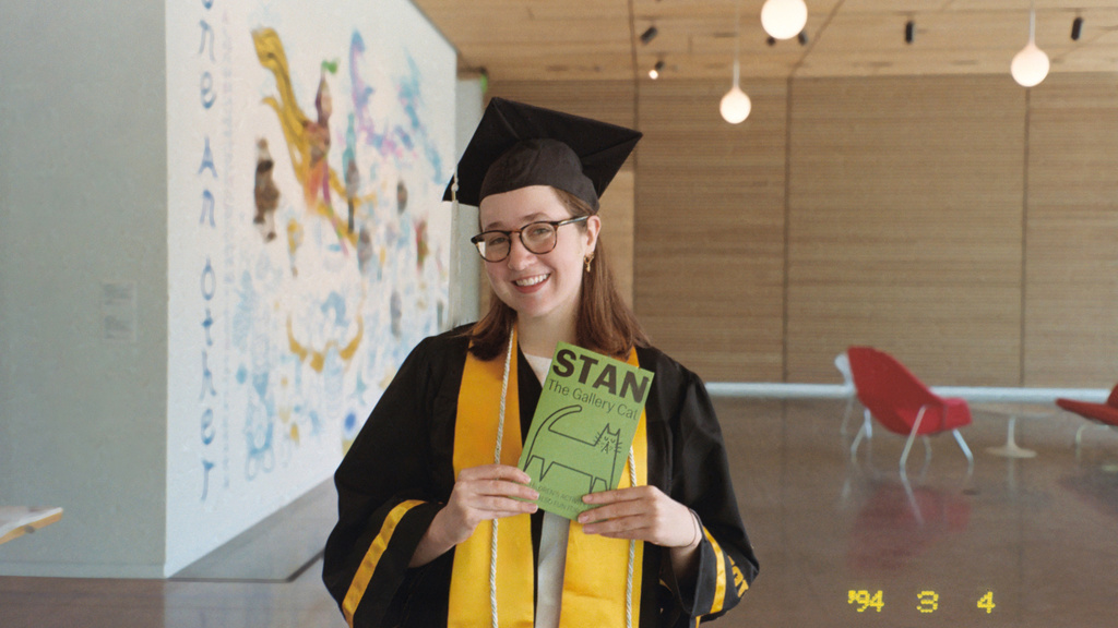 Museum Programs Intern Abbie McLaren, dressed in her graduation attire, poses with a Stan the Cat activity booklet, which she created in the spring of 2025, in the Stanley Museum of Art lobby.