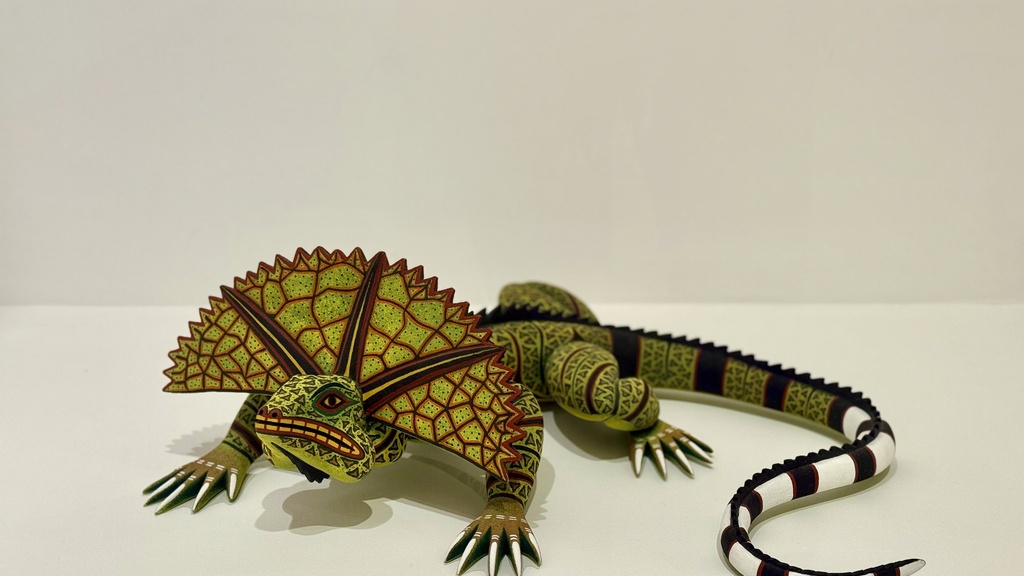 A sculpture of a green, intricately patterned lizard with a frilled neck covered in red geometric patterns and a black and white tail is displayed on a plain white surface.