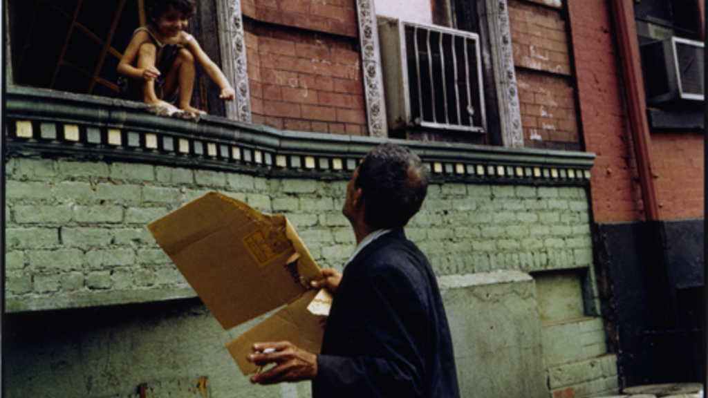 a man holding a broken cardboard box and a cigarette looking up at a young boy perched in a window above a green painted brick wall