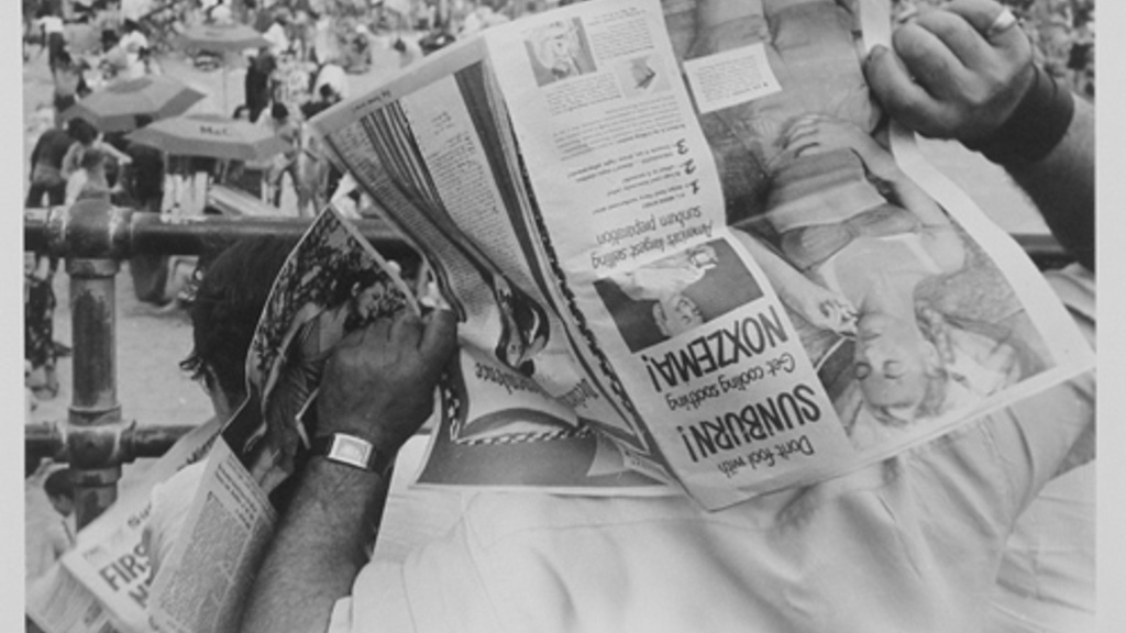 man holding an open newspaper over his head while he looks down at a beach crowded with people
