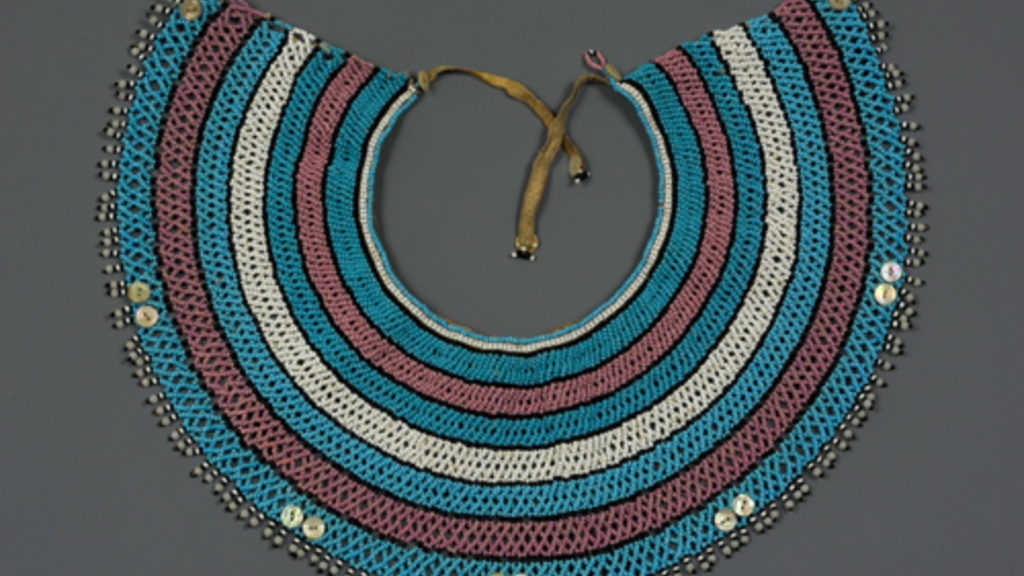 Wide beaded collar that forms most of a circle. The beading is intricate, and arranged colored bands from the smallest to largest: blue, pink, blue, white, blue, pink, blue with five pairs of white dots at regular intervals.