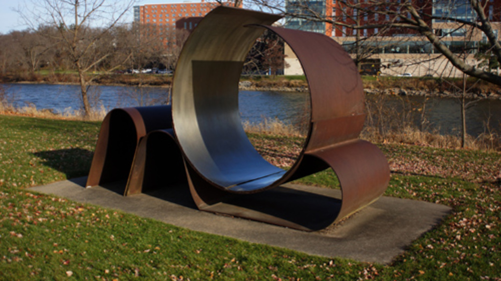 A large outdoor stainless-steel sculpture with wide flat pieces that form a wave shape below and curl into a near circle on top.