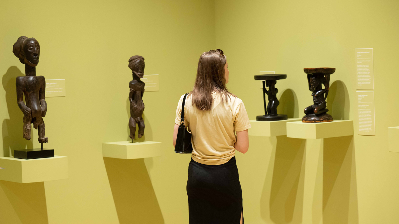 A guest stands in the corner of the "Fragments of the Canon" gallery: the walls are green, and they are surrounded by four works of art, carved from wood, on pedestals jutting out from the green painted walls.