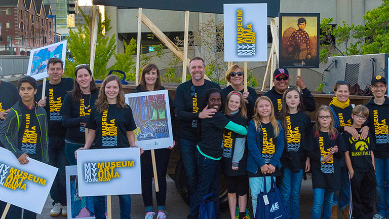 Group of people standing outside. They are wearing matching t-shirts and holding signs in preparation for the UI Homecoming parade.