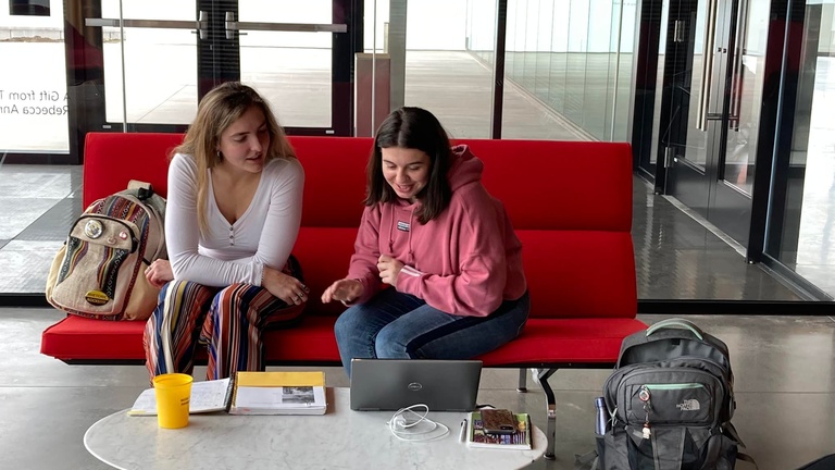 Two women sit on a red couch leaning over a white table. There is a laptop and beverage on the table and backpacks on the couch and floor.