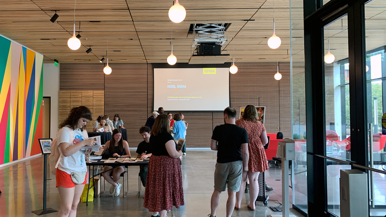 A photo taken at the HBD, SMA event. It shows one student in the foreground, looking at the HBD, SMA zine, and others in the back interacting with the gallery hosts at the event's information desk and mingling in the back.