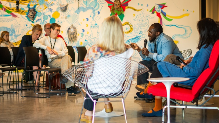 A man seated on a red couch holding a microphone speaks as a group of seated people listen