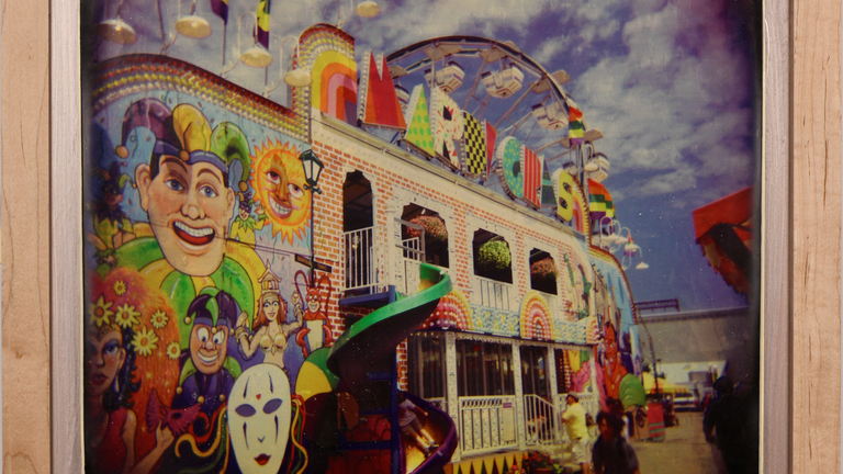 A two-story red brick building against a bright blue sky with fluffy white clouds. There is a colorful carnival themed mural painted on the front and a spiral slide from the top floor to the ground. A portion of a ferric wheel peeps out from behind the building.