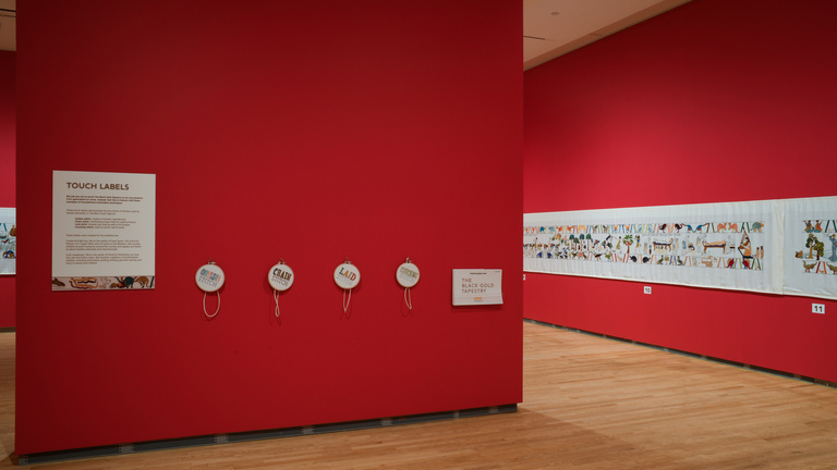 A red-wall in a gallery displays samples of embroidery with "The Black Gold Tapestry" by Sandra Sawatzky on view in the background.